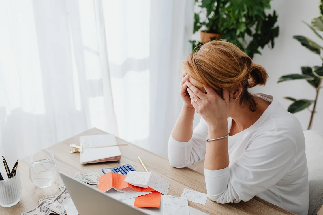 A woman at a home desk with her head in her hands, surrounded by receipts, sticky notes, and a calculator.